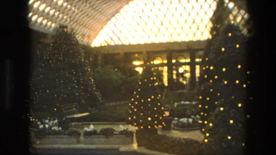 1972 Iran frame showing holiday-lit trees, low hedges, benches, and arched windows below the bright diamond-patterned glass awning.