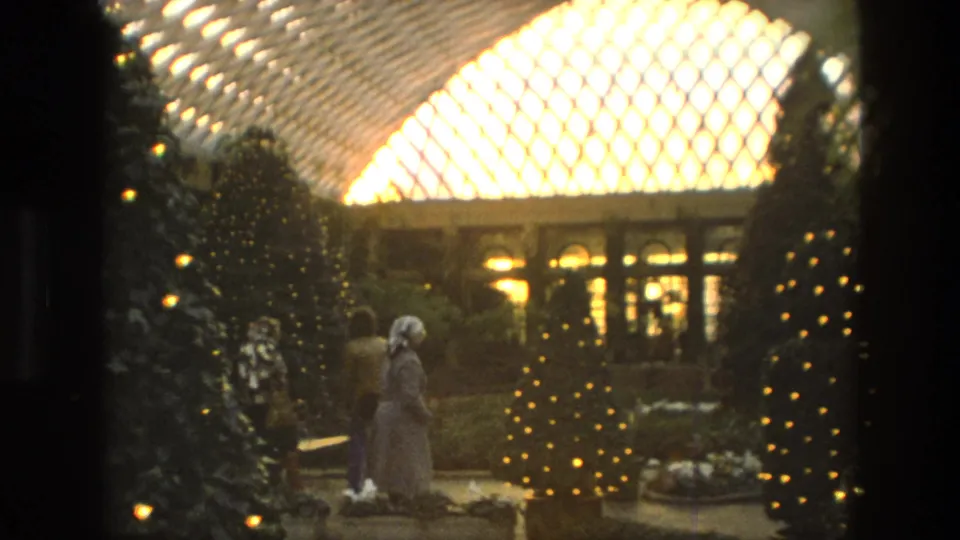 1972 Iran 8mm frame showing visitors near illuminated Christmas trees, indoor planting, a walkway, and a bright arched glass ceiling.