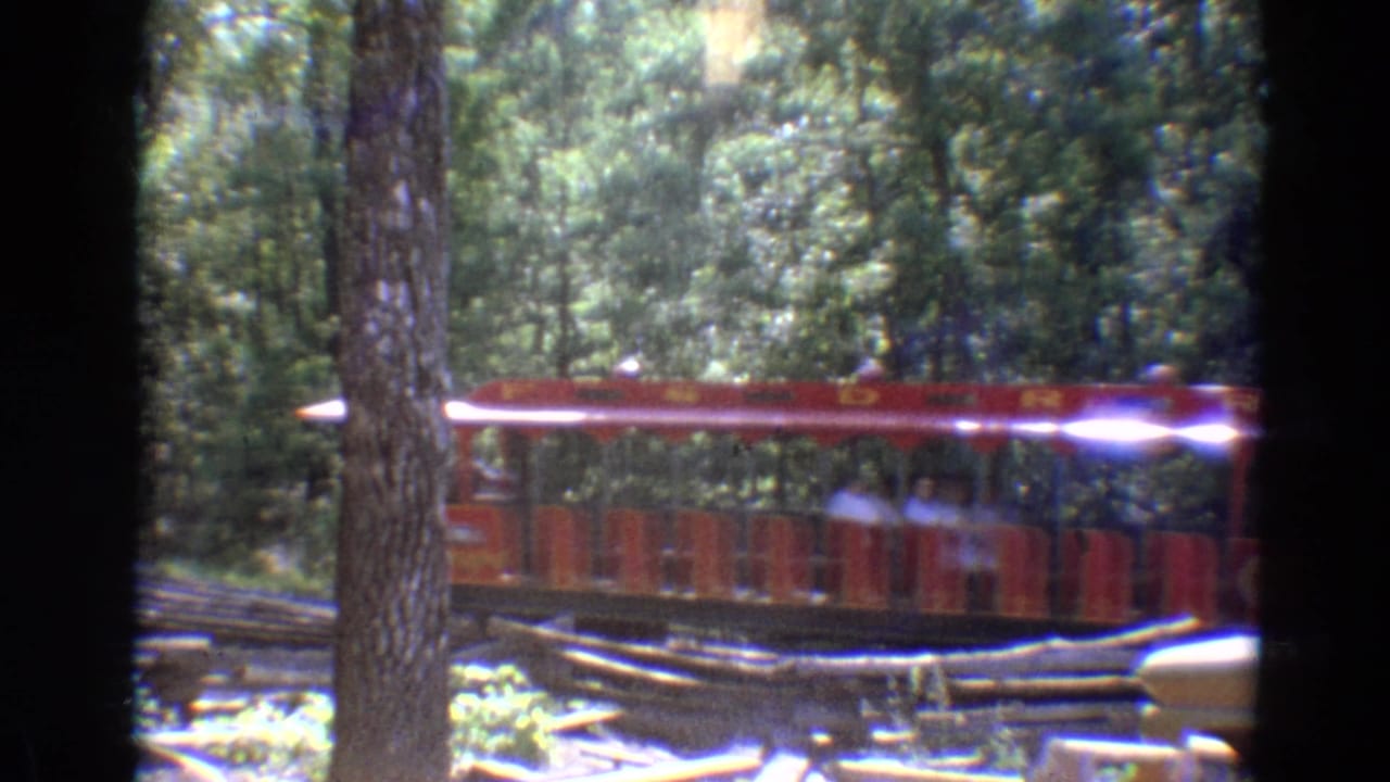 1968 Branson footage frame with red trolley cars and seated passengers moving through a wooded Silver Dollar City setting.