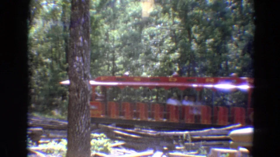 1968 Branson footage frame with red trolley cars and seated passengers moving through a wooded Silver Dollar City setting.