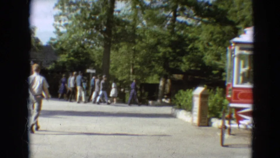 A group of visitors walks away along a broad path in the 1968 Branson home movie, with trees and a red vehicle or booth at the edge.