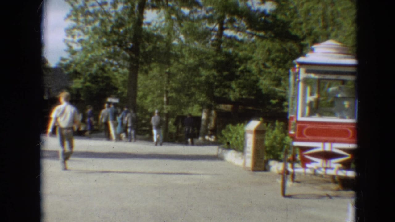1968 Branson fairground view with visitors on a paved path, trees overhead, and a red and white popcorn cart near the walkway.