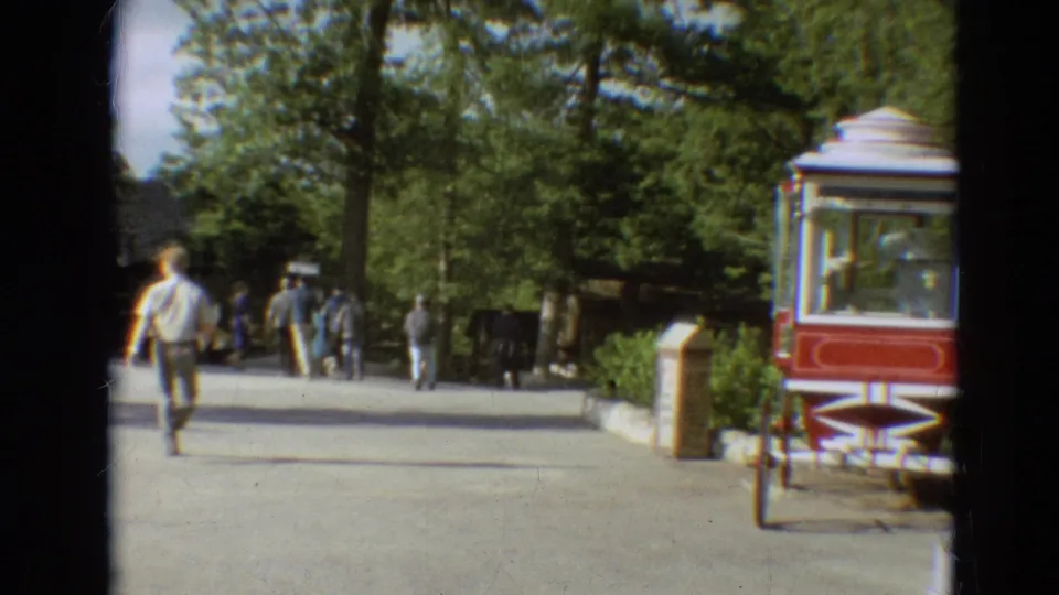 1968 Branson fairground view with visitors on a paved path, trees overhead, and a red and white popcorn cart near the walkway.