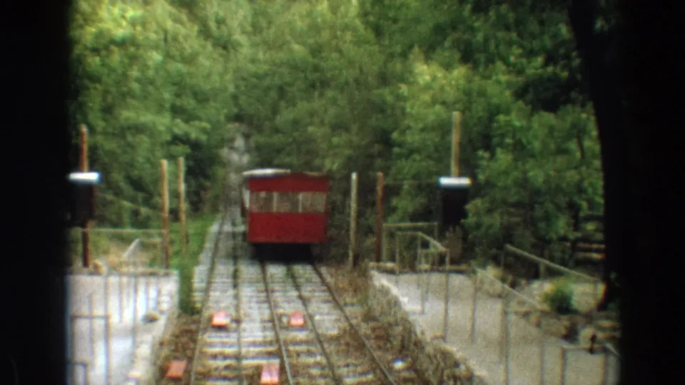 1968 Branson home-movie frame showing a red trolley moving along rails through trees, viewed from another trolley position.