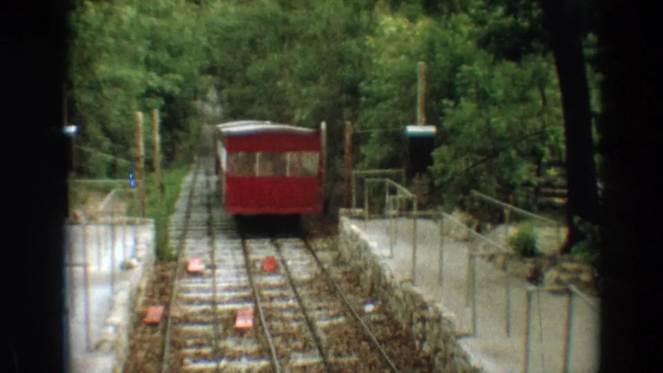 Vintage 8mm frame of a red Branson trolley nearing the camera on rails bordered by low walls, railings, and trees.