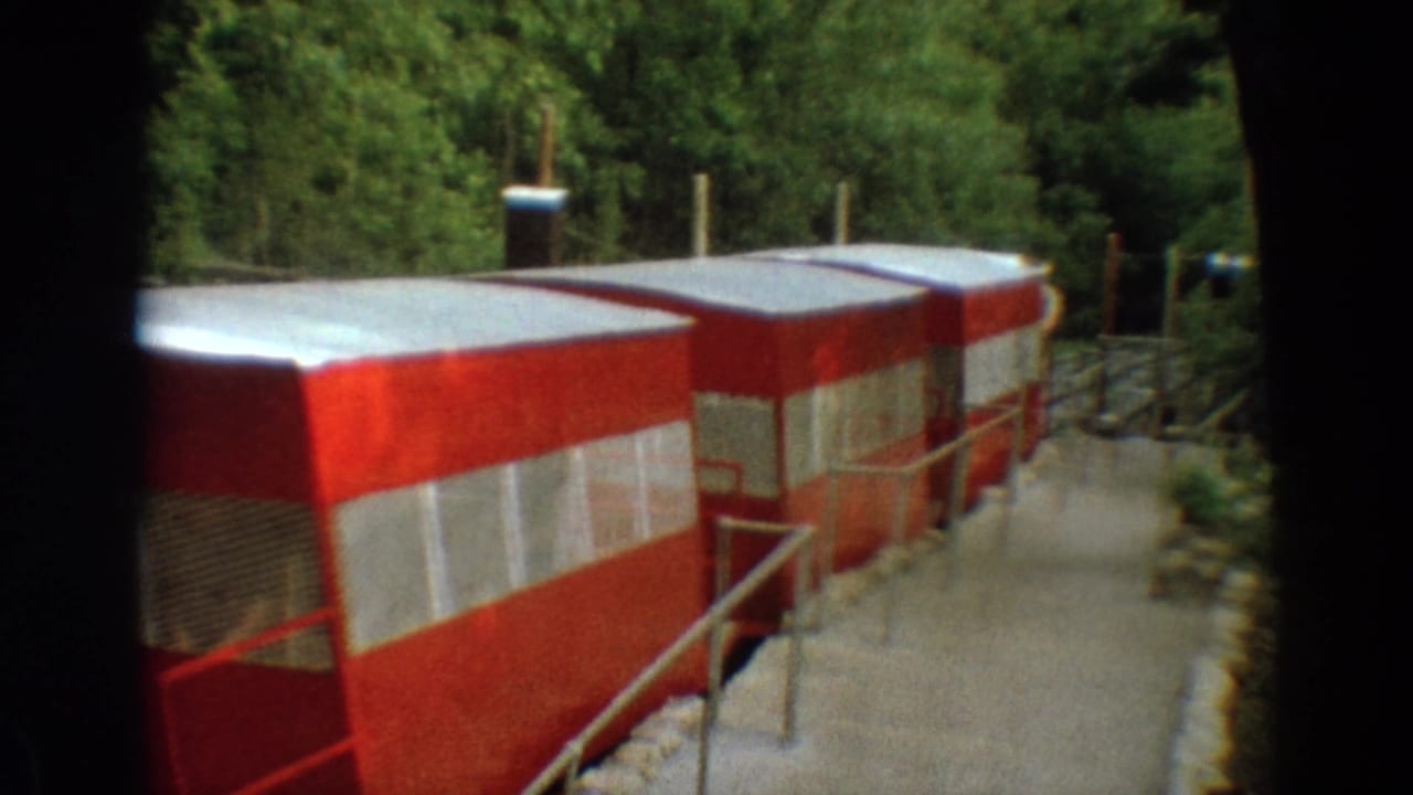 1968 Branson, Missouri home-movie frame showing red tram cars parked along a gravel walkway near dense trees.