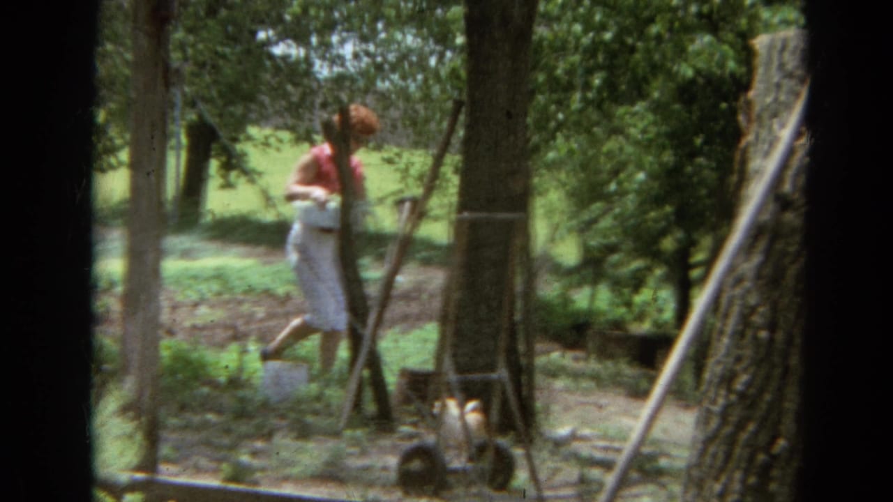 In 1968 Branson home-movie footage, a woman carries a white container through a shaded garden area near trees and a wire fence.