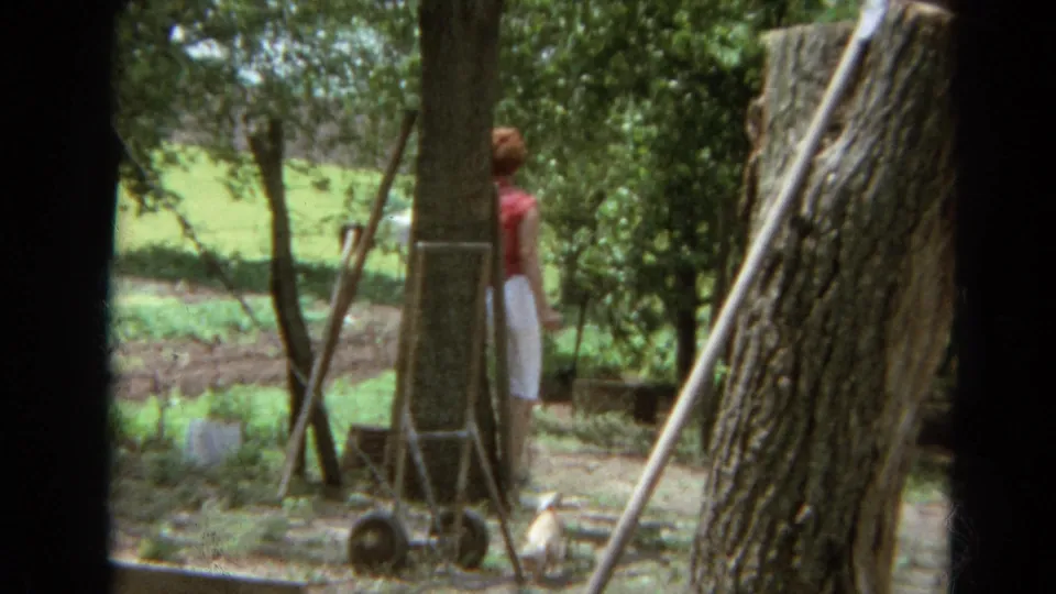 A woman in red and white stands near a tree, wire fence, and small cart in the 1968 Branson garden scene.