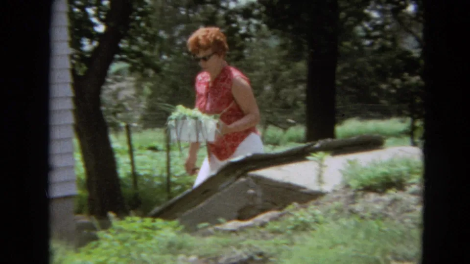 The woman continues across the garden near a white-sided house, holding a basket of green onions in 1968 Missouri home-movie footage.