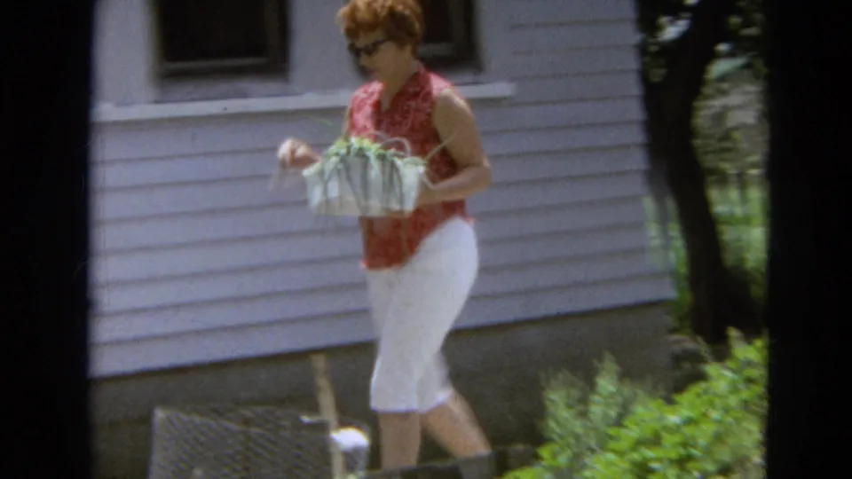 A woman in sunglasses and white capri pants passes a white-sided house while carrying a basket of green onions in Branson, Missouri.