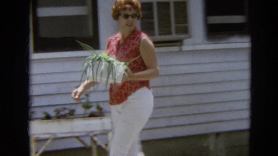 Near the white-sided house, the woman smiles toward the camera while holding green onions, with a window and yard furniture behind her.