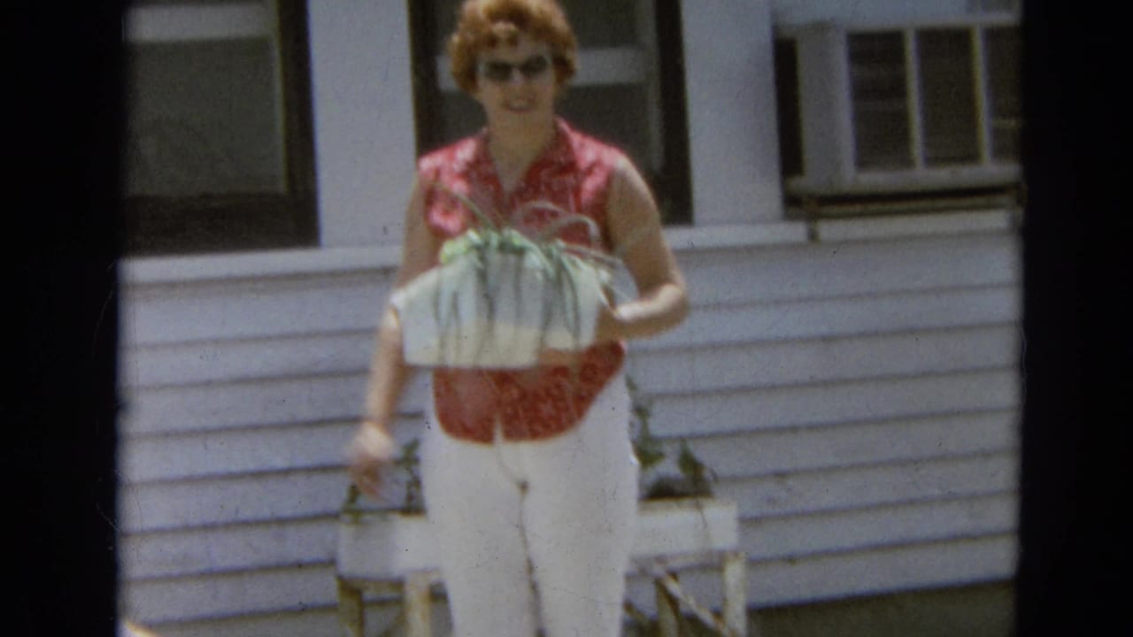 Woman in sunglasses carries green plants near a white house exterior.