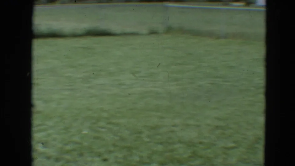 1968 8mm frame from Branson, Missouri showing an empty sunlit grass field bordered by a chain-link fence.