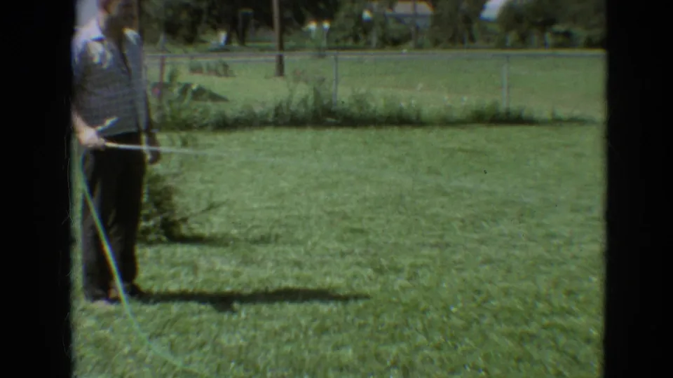 A man in a white shirt holds a hose on a Branson backyard lawn, with water spray crossing the 1968 home-movie frame.