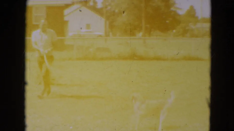 A bright, overexposed 8mm frame shows the man, hose, dog, lawn, fence, and house during the backyard play scene.