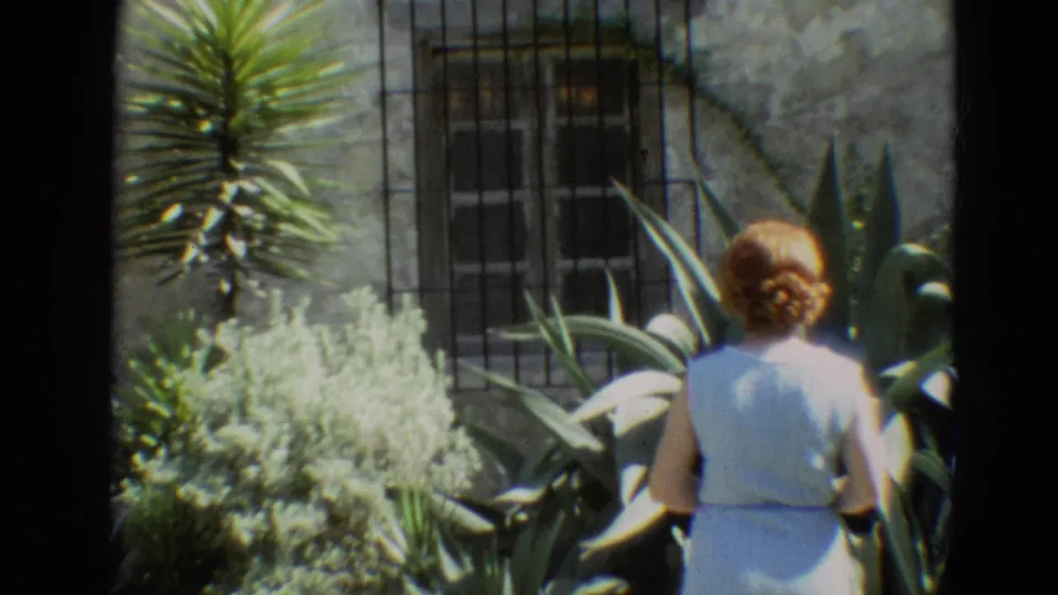A woman in a light blue dress walks away through the 1968 Branson garden, framed by succulents, pale shrubs, and a stone building.