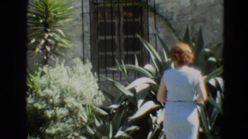 In 1968 Branson, Missouri footage, a woman in a white sleeveless dress stands among yucca-like plants and shrubs near a stone wall.