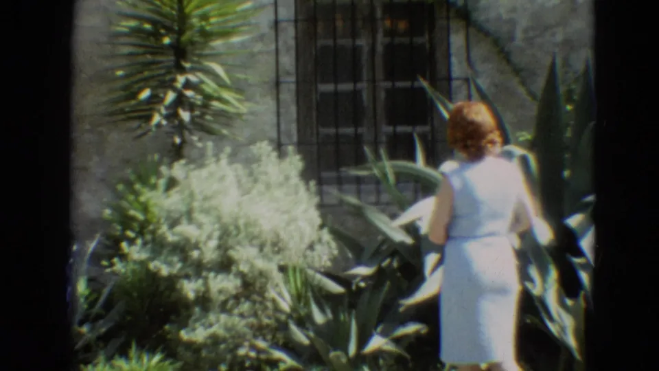 The woman moves through the Branson garden area, carrying a dark handbag near large succulents and a stone building wall.