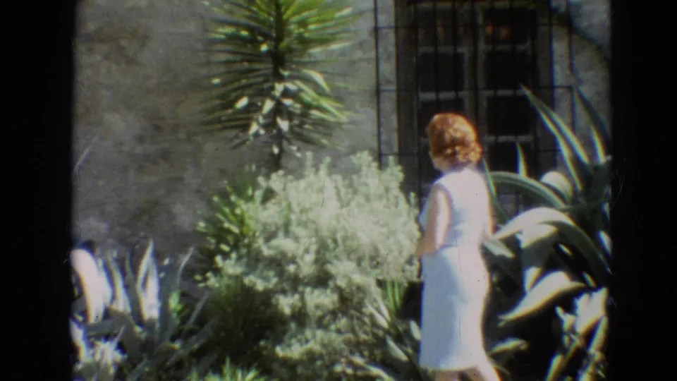 A side view shows the woman in a white dress beside pale shrubs and tall yucca-like plants in the 1968 Branson garden footage.