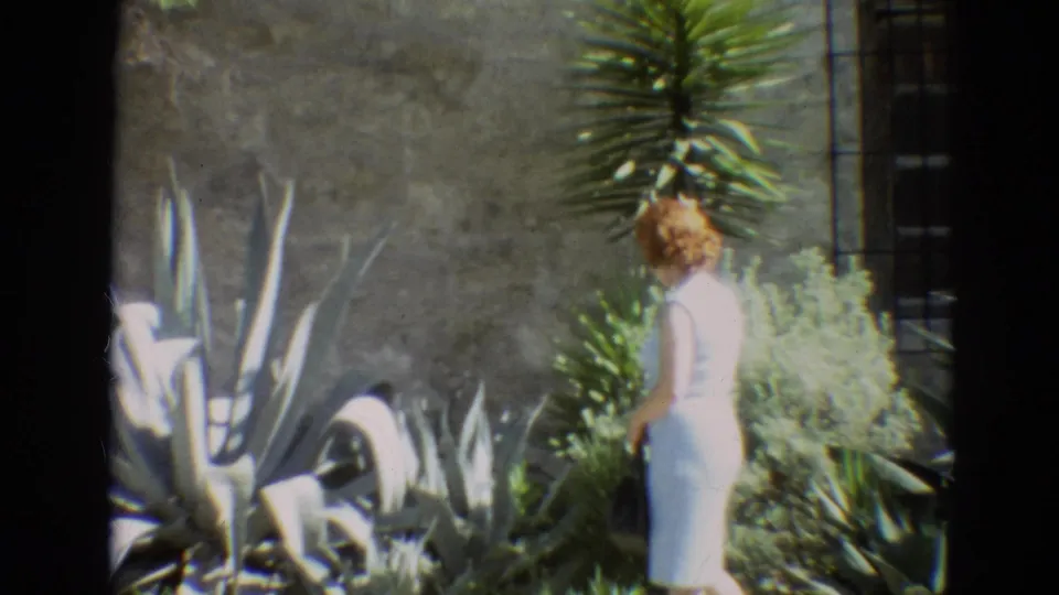 Near the end of the clip, the woman stands at frame right in the Branson garden, partly turned beside shrubs, succulents, and a stone wall.