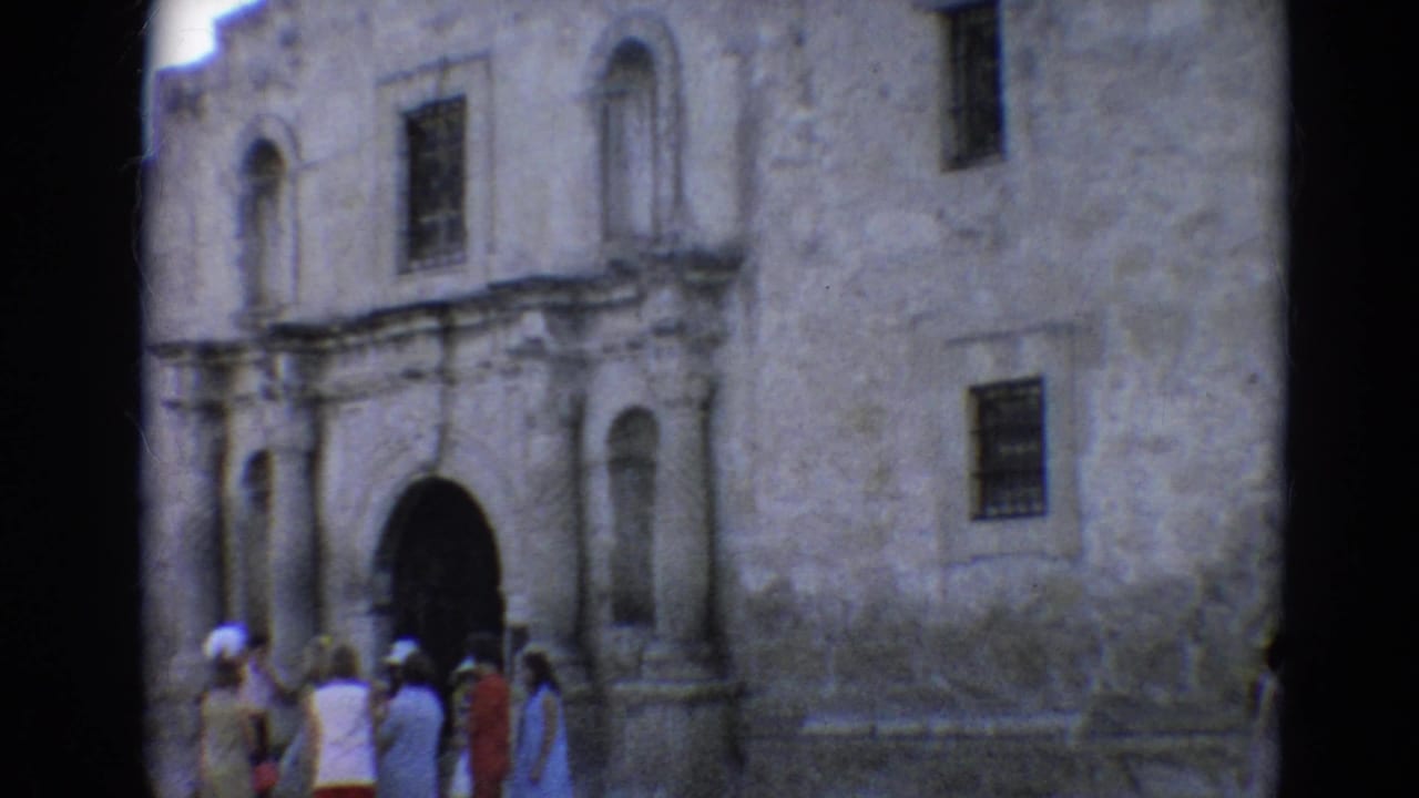 Tourists stand near a weathered stone building with arched entrances and barred windows.