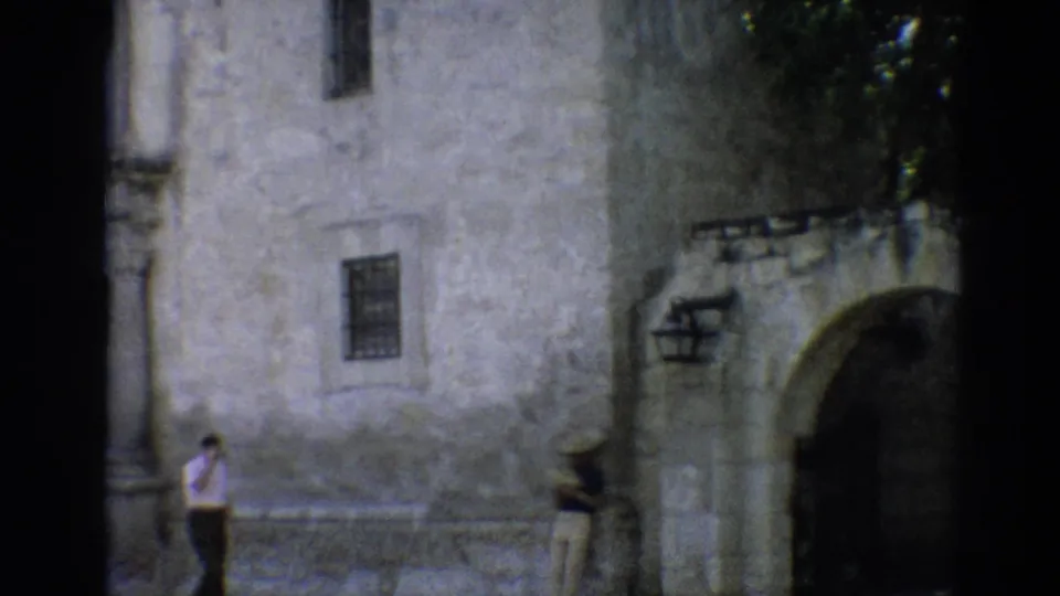 1968 Branson footage shows a man crossing the foreground beside a stone building and a person near a large shaded arch.