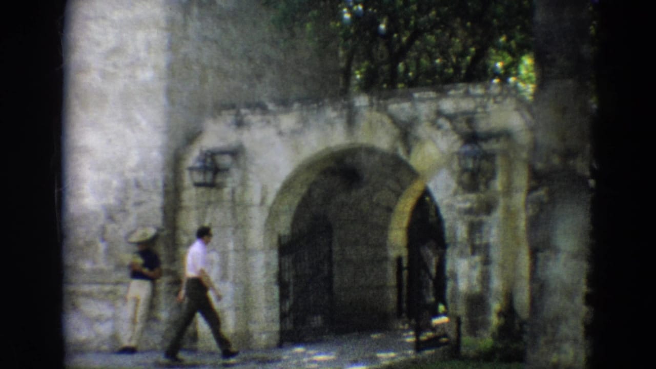 1968 Branson home-movie frame showing a man in a white shirt walking near a stone archway as another adult stands at left.