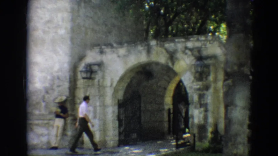 1968 Branson home-movie frame showing a man in a white shirt walking near a stone archway as another adult stands at left.