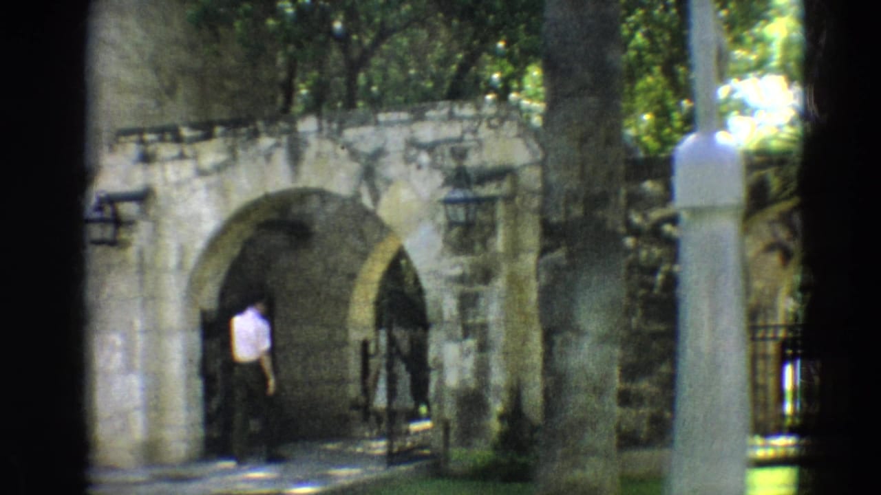1968 Branson frame showing a man in a white shirt beneath a weathered stone arch with trees and gatework nearby.