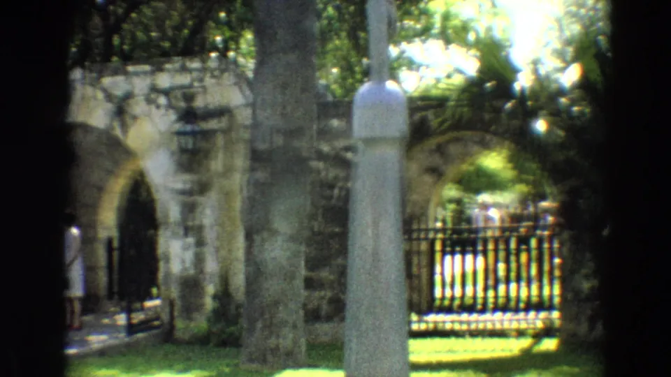 1968 Branson home-movie view of a stone arch, white post, iron fence, trees, and bright grass beyond.