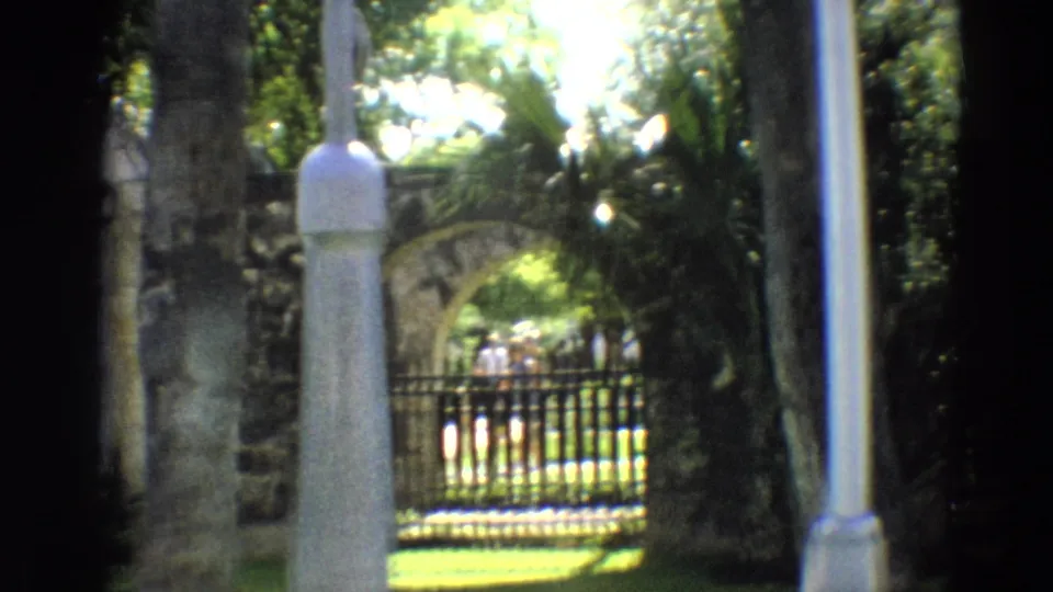 1968 Branson frame showing a white post, stone arch, iron gate, and shaded greenery in a historic outdoor setting.