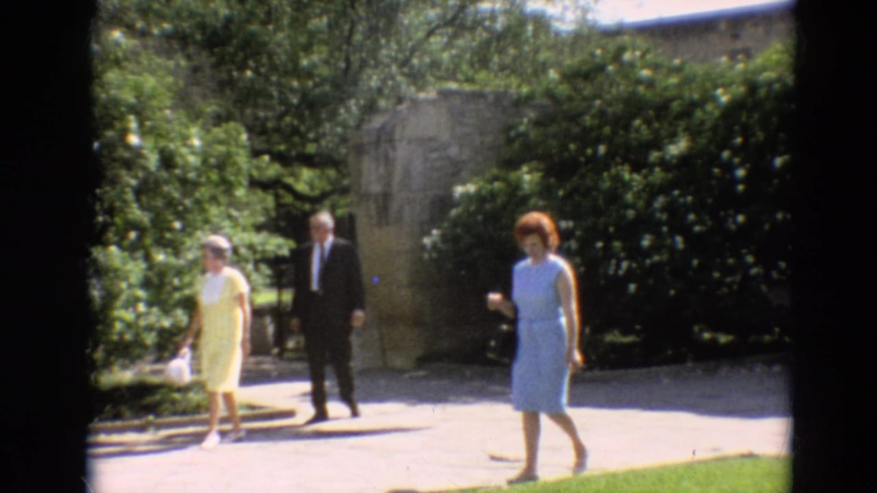 In metadata-supported 1968 travel footage, three adults walk outdoors along a landscaped paved path.