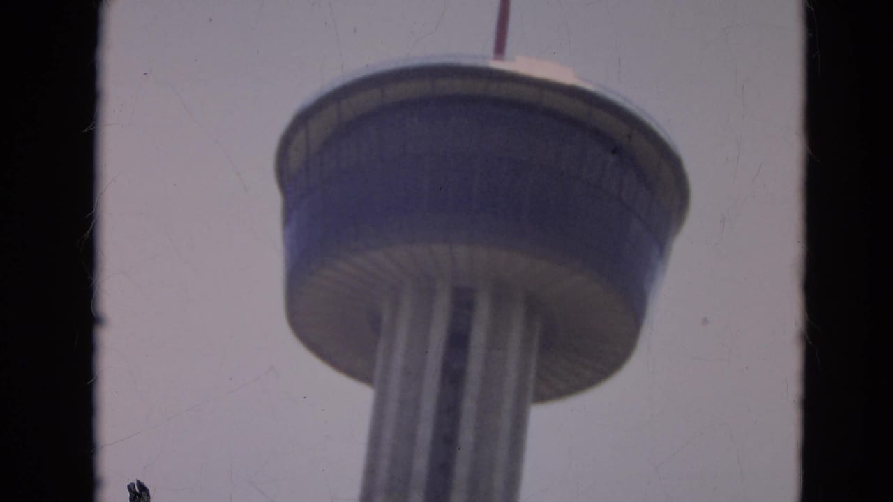 Low-angle 8mm view of the Seattle Space Needle observation deck and support columns against a pale sky.