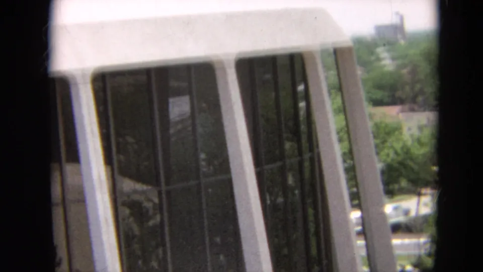 1968 Branson frame showing a pale windowed facade at an angle, with trees, cars, and distant buildings beyond.