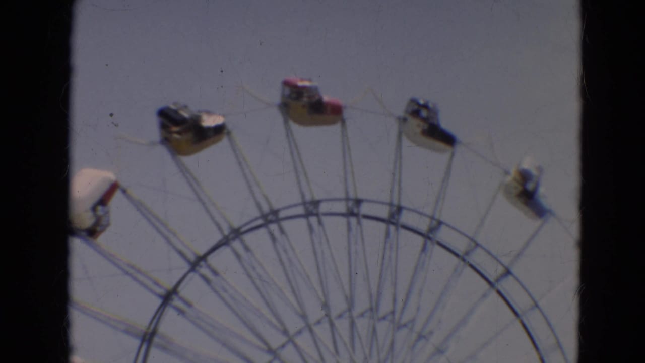 1968 Branson home-movie frame showing the upper arc of a Ferris wheel with small passenger cars against a pale sky.