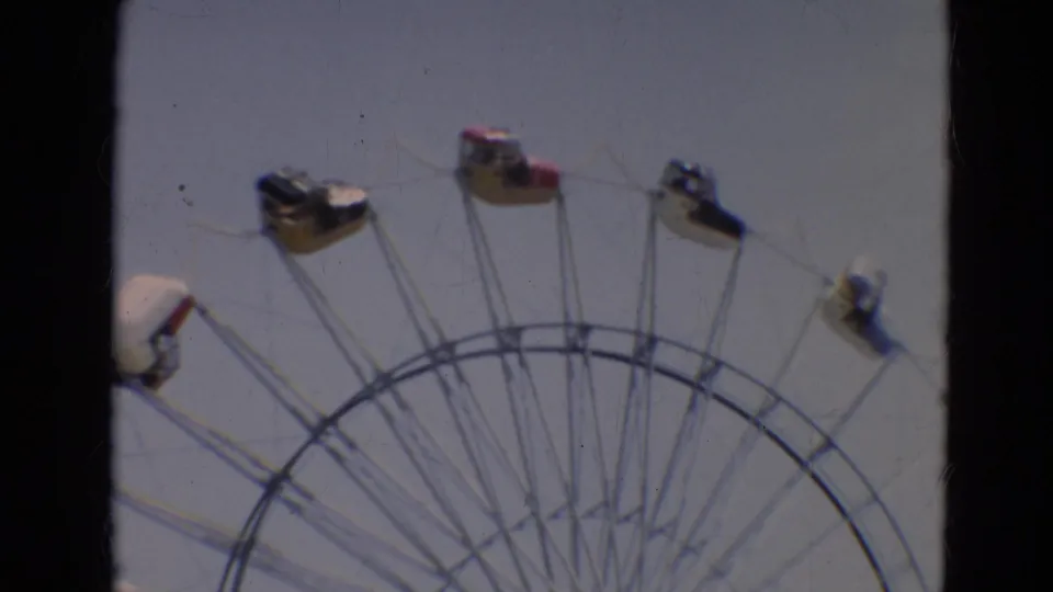 1968 Branson home-movie frame showing the upper arc of a Ferris wheel with small passenger cars against a pale sky.