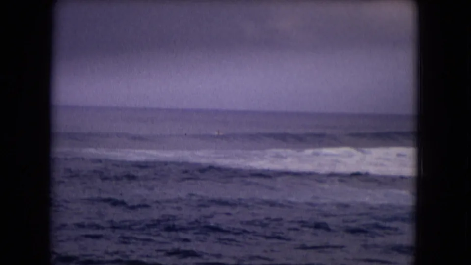 1968 Honolulu home-movie frame shows foamy waves moving across a dark shoreline view beneath a pale overcast sky.