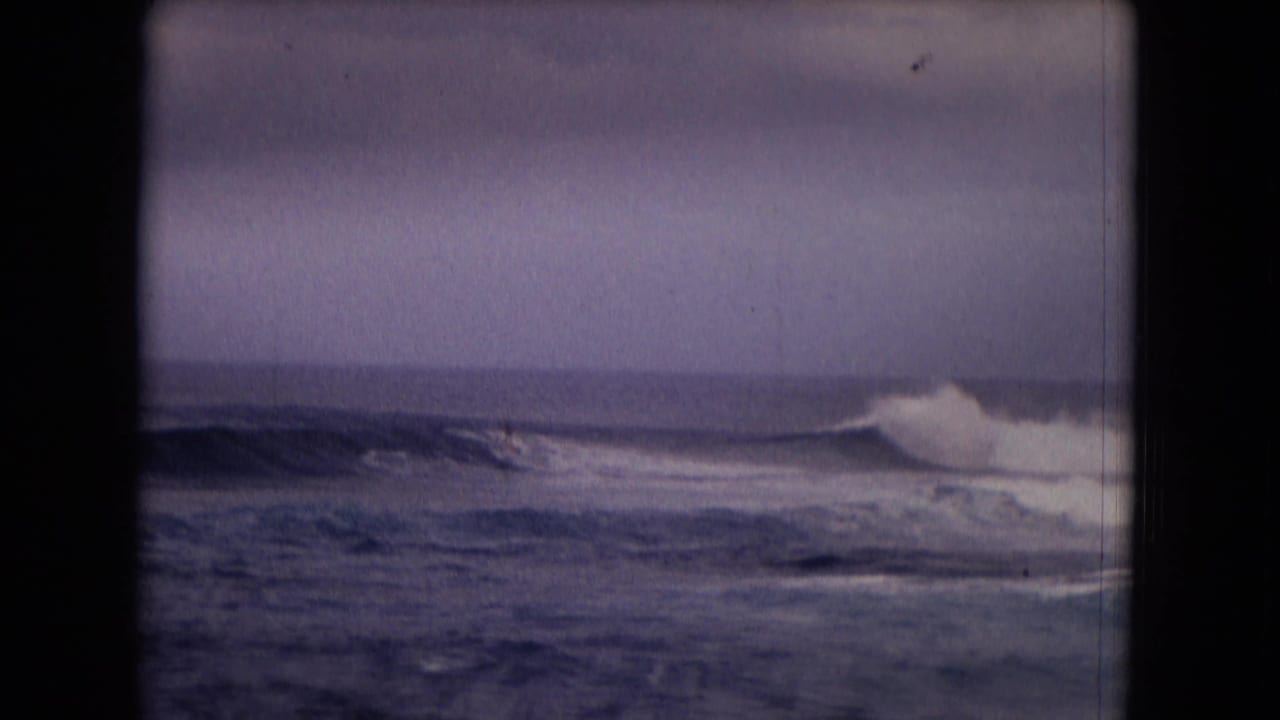 Breaking ocean waves beneath a muted gray sky, framed by dark 8mm film borders.