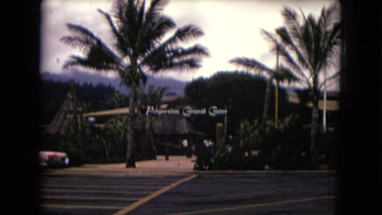 1968 Honolulu home-movie frame showing the Polynesian Cultural Center entrance area with palms, thatched roof, signage, and a parked car.