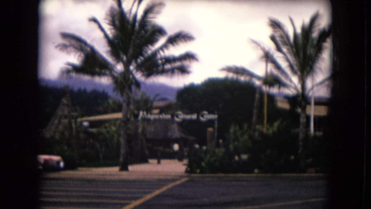 Entrance sign framed by palms, landscaped planting, a parked car, and distant mountains.