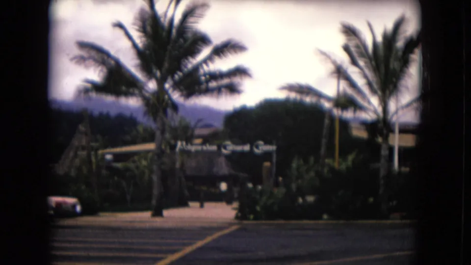 1968 Honolulu view of the Polynesian Cultural Center entrance sign, palms, tropical landscaping, parked car, and mountains beyond.