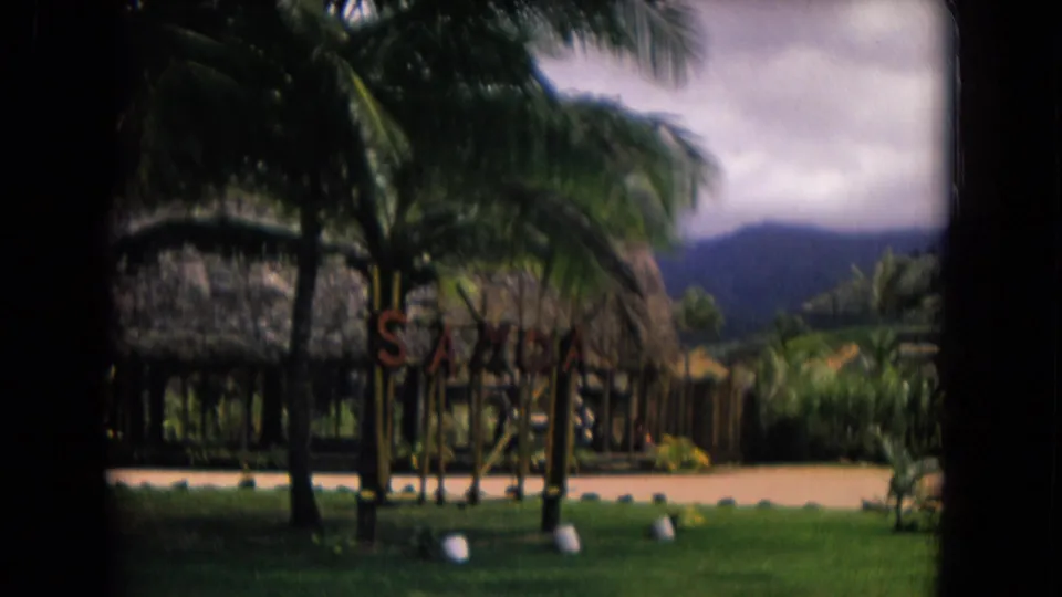 1968 Honolulu frame showing thatched structures and lawn near the Polynesian Cultural Center, with palms and mountains behind.