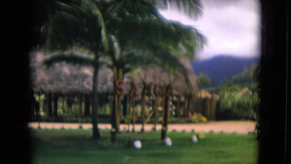 1968 Honolulu home-movie view across a grassy area toward thatched cultural-center structures, palms, fencing, and mountains.