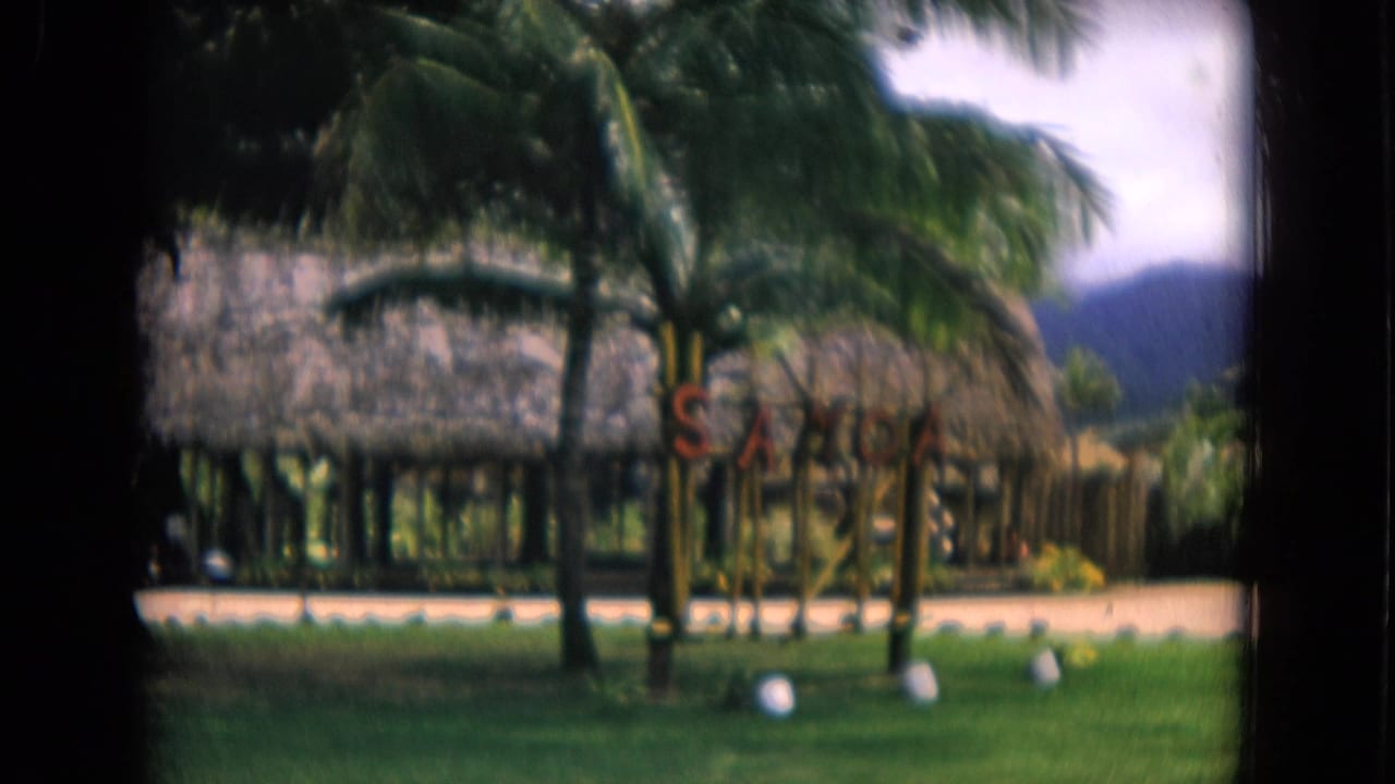1968 Honolulu home-movie view of thatched resort huts, palms, green lawn, small signage, and mountains beyond.