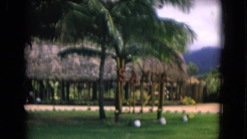 1968 Honolulu home-movie view of thatched resort huts, palms, green lawn, small signage, and mountains beyond.