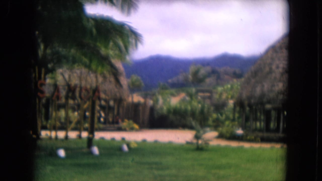 Honolulu resort grounds on 8mm film, showing palms, thatched structures, lawn, path, distant hills, and a small sign.