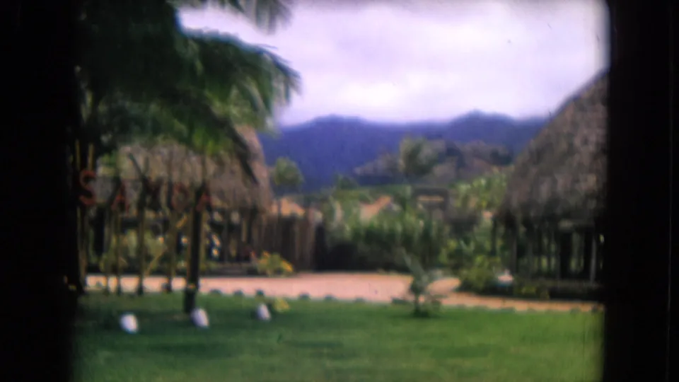 Honolulu resort grounds on 8mm film, showing palms, thatched structures, lawn, path, distant hills, and a small sign.