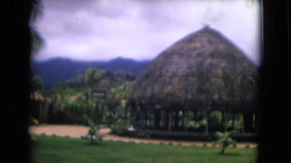 1968 Honolulu resort scene with a large thatched pavilion, tropical plantings, green lawn, path, and mountain backdrop.