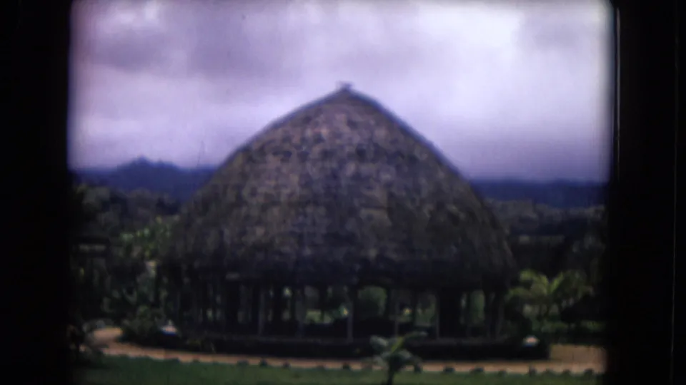 Honolulu home-movie frame of a round thatched pavilion set in resort landscaping with mountains under a cloudy sky.