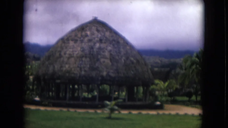 1968 Honolulu home-movie frame showing a thatched cabana, tropical landscaping, distant hills, and soft overcast sky.